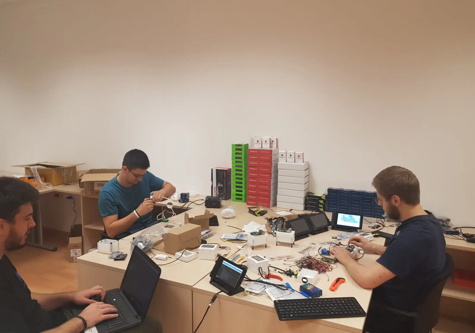 Three members of the META5 team at a long desk assembling hardware devices, with boxes of small touchscreen displays, cases, cables and a laptop